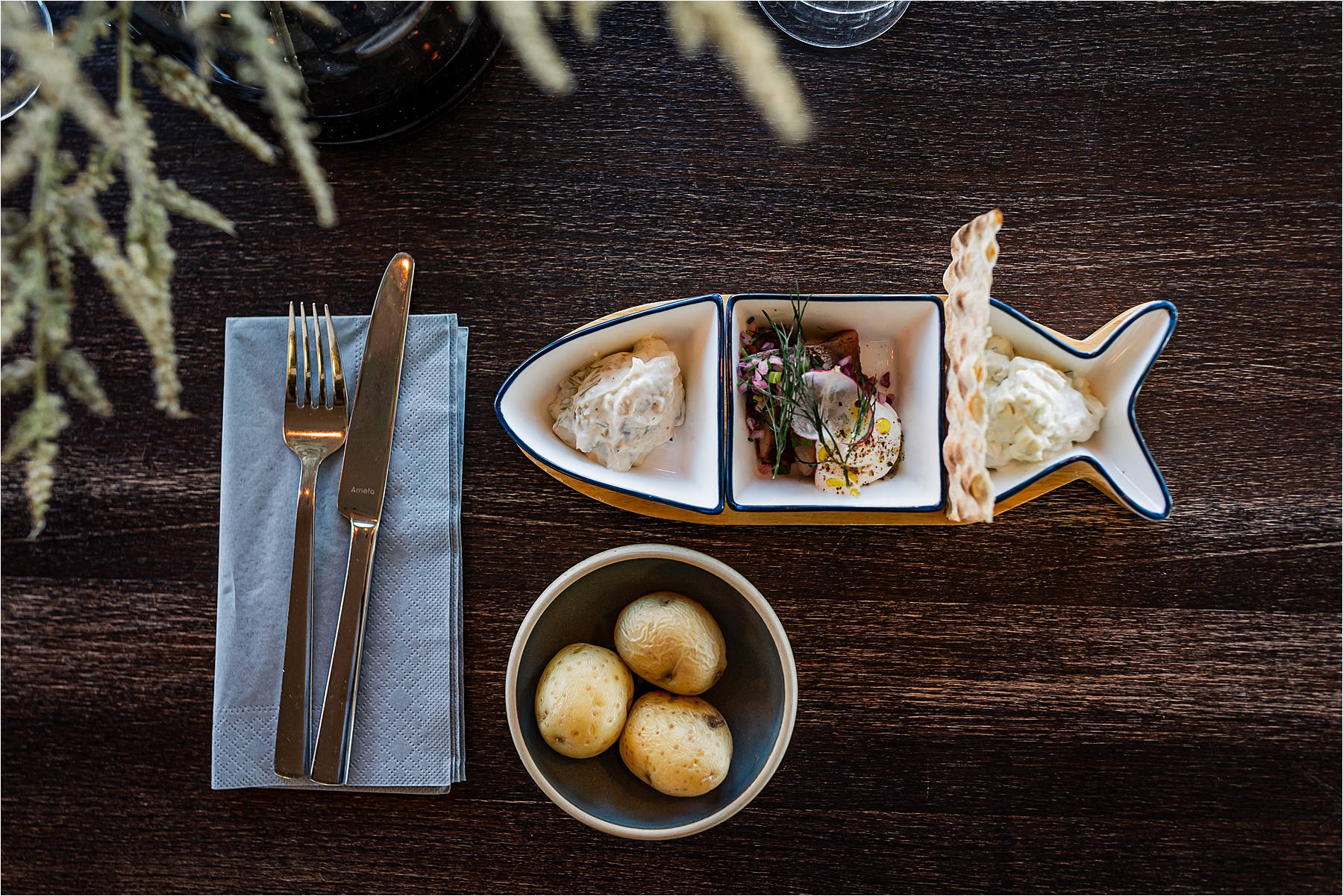 En man och en kvinna äter tillsammans på en fiskrestaurang på Smögenbryggan, sittande vid ett bord vid fönstret. De ler och är engagerade i samtal, med rätter, drinkar och blommor som pryder bordet. En annan kvinna syns i bakgrunden vid fönstret.